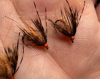 May include: Three artificial fishing flies featuring brown and black hackle feathers, tan bodies, and small bright orange beads at the hook eye. Each fly is constructed with thin, dark, pointed fibers extending outward from the central body.