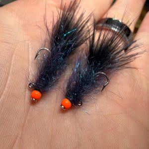 Puede incluir: Dos moscas de pesca con fibras negras, iridiscentes y azules. Cada mosca tiene una pequeña cuenta naranja en el ojal del anzuelo. Los anzuelos son plateados. Estas moscas están diseñadas para pescar.
