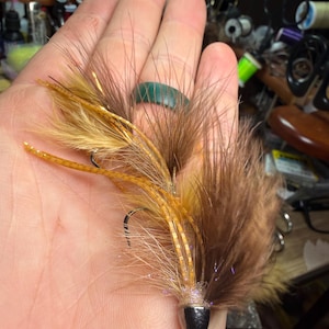 Puede incluir: Un señuelo de pesca con mosca hecho a mano con plumas marrones y doradas, un anzuelo negro y una cabeza plateada. La mosca se sostiene en una mano, mostrando los intrincados detalles. Este señuelo está diseñado para la pesca.