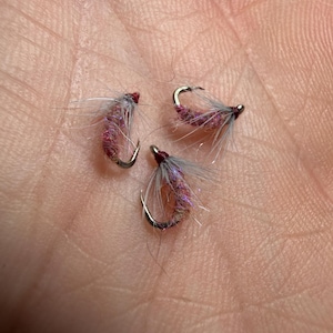Op de afbeelding: Drie kleine vliegvissen-aasjes met gebogen metalen haken, donkerrode lichamen met iriserende vezels en fijne grijze hackle-veren. Elke vlieg is geconstrueerd met een duidelijke kopwikkeling en delicate, pluizige synthetische materialen.