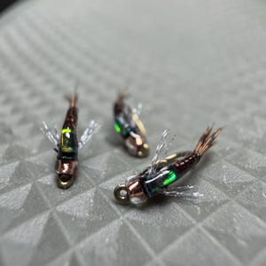 May include: Three artificial fishing flies. Each fly has a copper-colored body, iridescent green and blue accents, and a small hook. The flies also have small, white, feathery wings and brown tail feathers. The flies are arranged on a textured, gray surface.