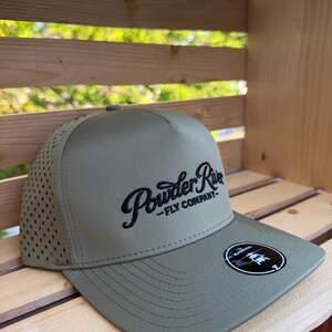 May include: An olive green baseball cap with a structured front panel and a perforated mesh back. The front features black embroidered text that reads Powder River FLY COMPANY. A circular black sticker is attached to the flat brim.