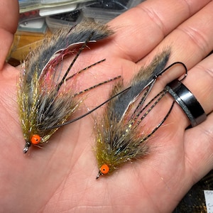 Puede incluir: Dos moscas de pesca hechas a mano. Cada mosca presenta un anzuelo, ojos de cuentas naranjas y un cuerpo de fibras sintéticas marrones, grises y doradas. Las moscas están diseñadas para imitar insectos y atraer peces.