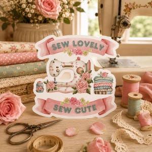 May include: A die-cut sign featuring a vintage sewing machine illustration, pink roses, and a stack of folded fabrics. The sign displays the text SEW LOVELY at the top and SEW CUTE at the bottom on pink banners, with a soft floral and craft theme.