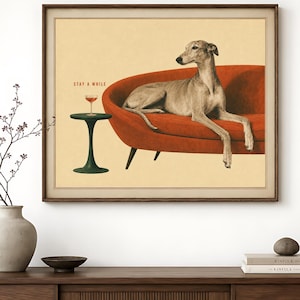 Pode incluir: Uma impressão de arte emoldurada apresentando um galgo descansando em um sofá laranja curvo ao lado de uma pequena mesa de pedestal verde com uma taça de coquetel. O texto STAY A WHILE está impresso em vermelho acima da taça em fundo bege.