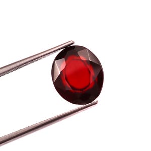 Puede incluir: Una gema facetada, de forma ovalada y color rojo intenso, es sostenida por unas pinzas plateadas sobre un fondo blanco. La gema tiene un corte brillante, que refleja la luz y muestra su rico color. Es probable que sea un granate.