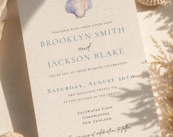 May include: A cream-colored wedding invitation card featuring a watercolor seashell illustration at the top. The text reads: Together with their loved ones, Brooklyn Smith and Jackson Blake invite you to their wedding celebration, Saturday, August 30th, two thousand twenty six, at five o'clock in the evening, Saltwater Cove, Coromandel, New Zealand. An evening of celebration to follow.