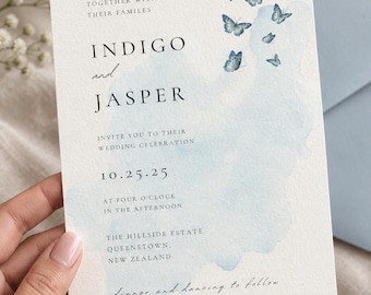 May include: A rectangular wedding invitation card on textured white paper featuring a light blue watercolor wash and small blue butterfly illustrations. The text reads: Together with their families, Indigo and Jasper, invite you to their wedding celebration, 10.25.25, at four o'clock in the afternoon, The Hillside Estate, Queenstown, New Zealand, dinner and dancing to follow.