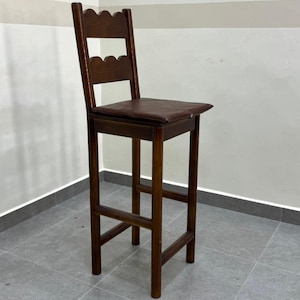 May include: A tall wooden bar stool with a dark brown finish, featuring a square seat with a dark brown padded cushion. The backrest has two horizontal slats with scalloped edges. The stool includes four straight legs connected by footrests.