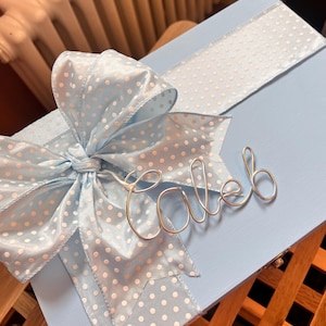 May include: A light blue gift box featuring a large satin bow and ribbon, both decorated with white polka dots. The name "Caleb" is written in silver wire on the box. The box is painted light blue.