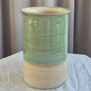 Könnte beinhalten: Eine zylindrische Keramikvase mit zweifarbigem Design, bestehend aus einer glänzenden hellgrünen Glasur im oberen Bereich und einem matten, unglasierten cremefarbenen Sockel. Das Gefäß hat einen leicht ausgestellten Rand und horizontale Texturlinien.