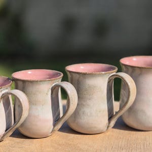 Op de afbeelding: Vier keramische mokken op een rij, elk met een afgerond lichaam, een lichtbeige buitenkant en een zachtroze geglazuurde binnenkant. Elke mok heeft een enkel gebogen handvat aan de zijkant en een consistent handgemaakt uiterlijk.