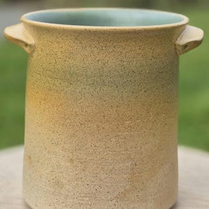 Op de afbeelding: Een cilindrische keramische pot met een gespikkelde beige en gele buitenkant die naar de bovenrand toe overgaat in een zachte groene tint. De pot is voorzien van twee kleine, uitstekende handvatten en een lichtblauwe geglazuurde binnenkant.