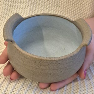 Peut inclure: Un petit bol en céramique beige moucheté, de forme ronde, doté de deux anses latérales intégrées. L'extérieur présente une finition texturée et terreuse, tandis que l'intérieur est recouvert d'un émail lisse bleu clair, créant un contraste.