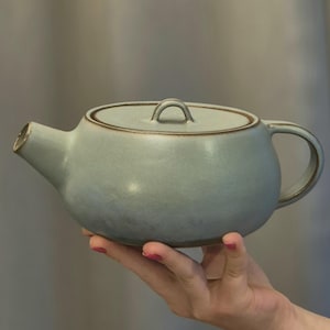 Op de afbeelding: Een lichtblauwe keramische theepot met een glad, rond lichaam, een korte tuit en een bijpassend deksel met een kleine boogvormige handgreep. De rand van het deksel, de opening van de tuit en de randen van het handvat hebben een bruine afwerking.