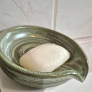 Op de afbeelding: Een glad, wit, rechthoekig stuk zeep ligt in een rond, saliegroen keramieken zeepbakje. Het bakje heeft een licht opstaande rand, een ondiepe kom met zichtbare cirkelvormige groeven en een kleine tuit aan één kant voor afwatering.