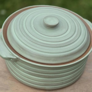 Op de afbeelding: Een ronde keramische ovenschaal met bijpassend deksel, voorzien van een licht saliegroen glazuur over een terracotta basis. De buitenkant en het deksel zijn versierd met horizontale concentrische ribbels en de schaal heeft twee handgrepen.