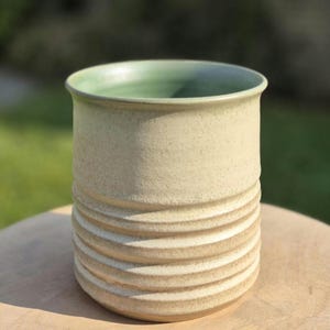 Op de afbeelding: Een cilindervormige keramische beker met een matte, crèmekleurige buitenkant met horizontale ribbels op de onderste helft. De binnenkant van de beker is geglazuurd in een gladde, licht saliegroene afwerking die contrasteert met het textuuroppervlak.