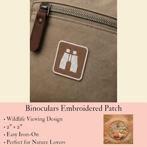 May include: A square-shaped embroidered patch featuring a white graphic of binoculars on a brown background. The patch is attached to a tan fabric surface with a visible zipper and a brown leather-like pull tab. The patch measures 2 inches by 2 inches.