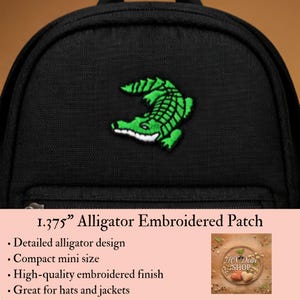 May include: A black backpack featuring a green alligator embroidered patch. The patch measures 1.375 inches and displays a detailed alligator design. The backpack has a textured fabric surface and a zippered pocket located below the patch.