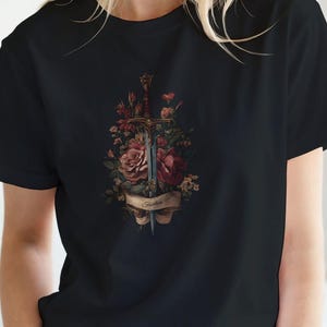 Può includere: T-shirt nero con un design floreale e una spada. La grafica include rose rosse e rosa, una spada e la parola "Justice" su uno stendardo. La maglietta è in stile girocollo classico.