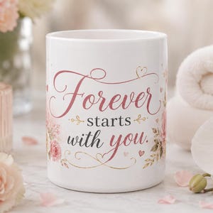May include: A white ceramic mug featuring the text Forever starts with you in elegant pink and dark gray script lettering. The design is decorated with delicate pink floral illustrations and small heart accents on both sides of the central text.