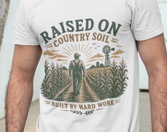 May include: White t-shirt with a graphic featuring a farmer walking through a cornfield, a windmill, and the text "RAISED ON COUNTRY SOIL - BUILT BY HARD WORK." The shirt is worn by a person wearing a gray baseball cap.