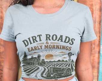 May include: A light gray crop top with the text "DIRT ROADS & EARLY MORNINGS" above a graphic of a farm scene. The graphic includes a farmhouse, tractor, and windmill. The model is wearing light blue denim shorts.