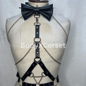 Op de afbeelding: Zwart leren harnas met een vlinderdas-kraag met zilveren kettingaccenten en metalen ringen. De vlinderdas heeft zilveren spikes. Op het harnas staat de tekst "BodyXCorset".