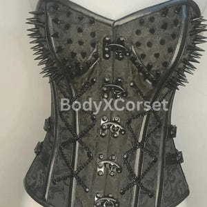 Può includere: Un corsetto nero con un motivo floreale, adornato con chiusure metalliche, catene e borchie appuntite sul bordo superiore. Il testo "BodyXCorse" è visibile. Il corsetto ha un design strutturato e aderente.