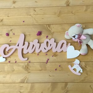 Pode incluir: Uma placa de madeira rosa com a palavra "Aurora", acompanhada de um urso de pelúcia branco de crochê com um laço rosa, um coração branco e um cavalo de baloiço rosa. Os itens estão dispostos sobre uma superfície de madeira castanha clara.