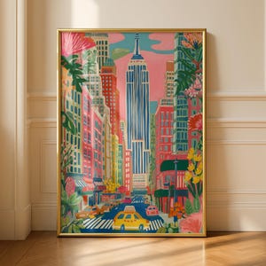 May include: A framed vertical art print depicting a New York City street scene with the Empire State Building centered. The illustration features vibrant pink, blue, and yellow tones, with stylized buildings, a yellow taxi, and lush floral foliage.
