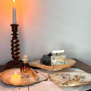 May include: A tall wooden spiral candlestick with a lit white taper candle, two irregular shell-shaped dishes, a perfume bottle, a gold lipstick tube, a Dolce & Gabbana perfume bottle, a black box, a small tea light candle, and silver jewellery.