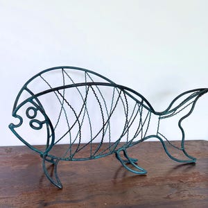 May include: A decorative fish-shaped rack constructed from dark teal wire with a weathered, oxidised finish. The structure features a curved spine, a circular eye detail, and a wide, open-frame body design supported by two thin wire legs.