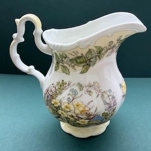 Puede incluir: Una jarra de leche de porcelana blanca con un asa decorativa y un borde festoneado. El cuerpo está adornado con una colorida ilustración de flores de primavera amarillas, follaje verde y un pequeño ratón entre las plantas del jardín.