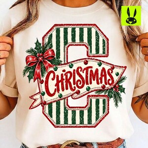 May include: A cream-coloured t-shirt featuring a large letter C with green and white vertical stripes. A banner across the letter reads CHRISTMAS in red script, decorated with green holly leaves, red berries, and a red bow on the left side.
