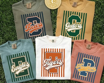 May include: Five crew neck t-shirts in olive, tan, light blue, white, and coral, each featuring a large vertical striped square, a bold block letter, and a script team name: Bears, Gators, Eagles, Tigers, and Panthers. A circular logo reads Timeless Craft.