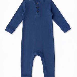 Può includere: Un body blu per neonati, con maniche lunghe e trama a coste. Presenta uno scollo rotondo e tre bottoni marroni. I polsini e le caviglie sono blu tinta unita.