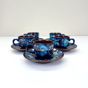 May include: A set of four dark blue ceramic cups and matching saucers, each featuring a hand-painted floral motif in shades of light blue, white, and yellow. The cups have a rounded handle and a smooth, glossy finish, arranged in a group.
