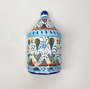 Könnte beinhalten: Eine wandmontierte Keramik-Laterne mit konischer Oberseite und zylindrischem Körper mit herzförmigen Ausschnitten. Das Stück ist handbemalt mit einem bunten Blumen- und Vogelmotiv in Blau-, Gelb-, Grün- und Rottönen auf weißem Untergrund.