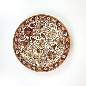 Peut inclure: Une assiette ronde en céramique ornée d'un motif floral peint à la main dans des tons de brun, de beige et de blanc. Le centre est rempli de diverses fleurs, tandis que la bordure présente un motif répétitif de petites fleurs blanches.