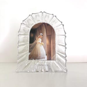 May include: Clear, arched photo frame with a rippled, textured border. The frame holds a photograph of a bride in a white wedding dress, holding a bouquet. The frame is displayed on a white surface.