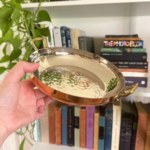 May include: A round, shallow copper pan with a polished, reflective interior surface and two small, curved brass handles on opposite sides. The exterior of the pan features a warm, metallic copper finish with a smooth, rounded rim.