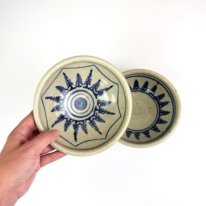 May include: Two round ceramic bowls with a speckled light beige finish. The interior of each bowl features a dark blue hand-painted design consisting of a central circle surrounded by a radial pattern of stylised evergreen tree silhouettes.