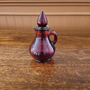 May include: A small, deep red glass cruet featuring a rounded body, a curved handle on the side, and a matching pointed glass stopper. The vessel has a smooth, translucent finish and a narrow neck, designed for pouring liquids with a small spout.