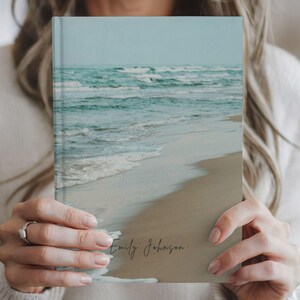 May include: A hardcover journal featuring a serene beach scene with turquoise water and a sandy shore. The cover displays the name "Emily Johnson" in elegant script. The journal is held by a person.