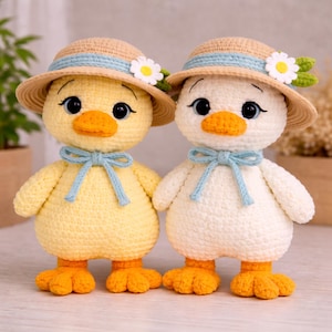May include: Two handmade crochet ducklings. One is yellow, the other is white. Both wear straw hats with blue bands and daisy accents, and blue bow ties. They have orange beaks and feet, and black eyes.