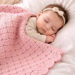 May include: A sleeping baby wrapped in a soft, pink crocheted blanket. The blanket has a textured square pattern and a scalloped edge. The baby wears a headband with a white bow.