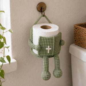 Op de afbeelding: Een handgemaakte, groene gehaakte toiletpapierhouder in de vorm van een figuur, met een wit kruis op de voorkant. De houder hangt aan een messing haak en houdt een rol wit toiletpapier vast. Het ontwerp omvat armen, benen en een lus om op te hangen.