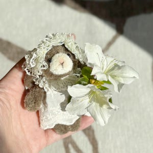 Könnte beinhalten: Eine kleine, flauschige braune Teddybär-Figur, die eine weiße Spitzenhaube und ein passendes weißes Spitzenkleid trägt. Der Bär hält ein Bündel aus drei weißen Blüten mit grünen Blättern, die zarte Blütenblätter und eine weiche Textur aufweisen.